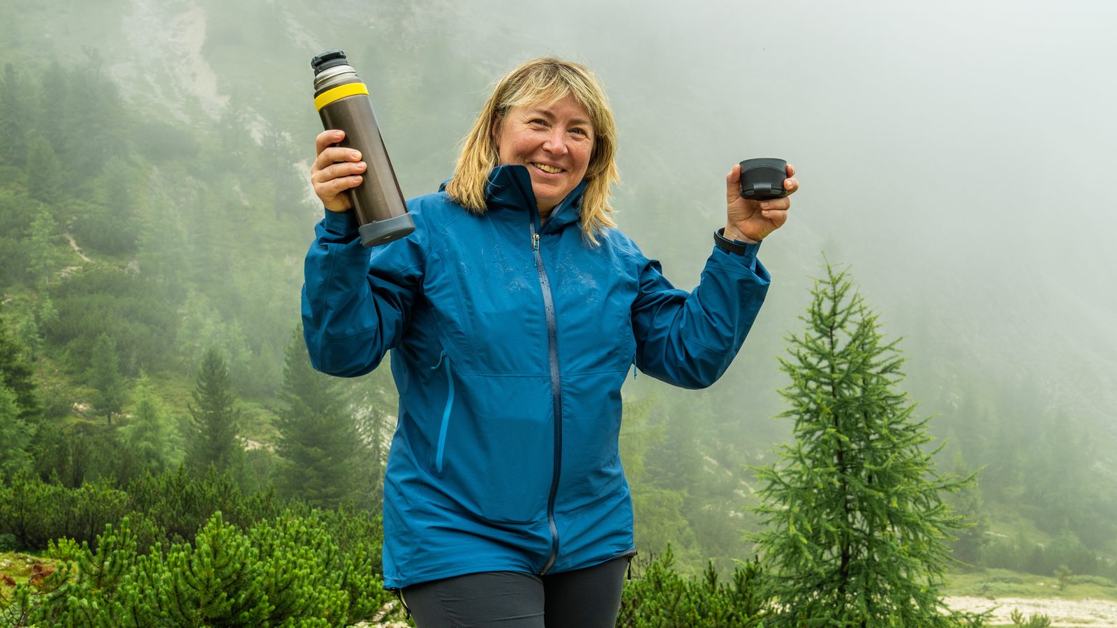 A walker in a waterproof jacket holding a flask on a misty forested slope